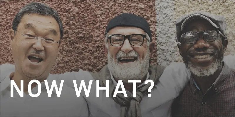 3 older men of different nationalities stand together with arms around each other. All men are smiling and are in their senior years.